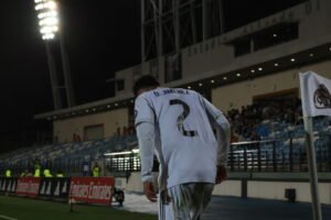 Jugador de fútbol con camiseta blanca número 2 caminando cerca del terreno de juego durante un partido nocturno.